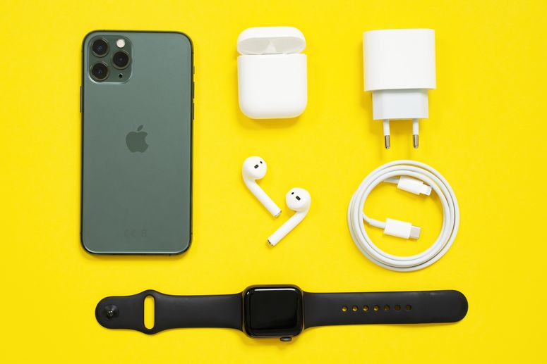 Conjunto de dispositivos de Apple: un iPhone negro y un Apple Watch, unos AirPods blancos con su caja, un cargador y un cable Lightning de Apple a USB tipo C, dispuestos en filas ordenadas sobre un fondo amarillo.